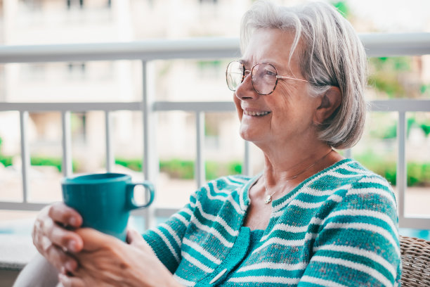 老妇人坐在家里的阳台上，端着咖啡杯享受片刻的放松，戴着眼镜的白发老妇人微笑着，心情很好图片下载