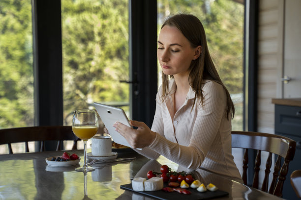 女人穿着家常的衣服，在厨房内部的背景下，坐在桌子旁，吃着水果、奶酪和一杯咖啡，使用数码平板电脑或浏览互联网图片下载