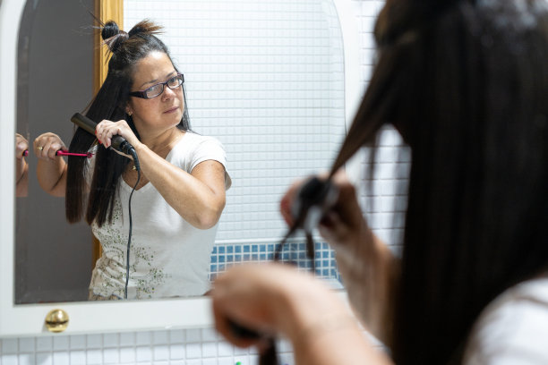 一个女人在家里用陶瓷熨斗烫直头发，看着浴室镜子里的自己。护发理念。图片下载