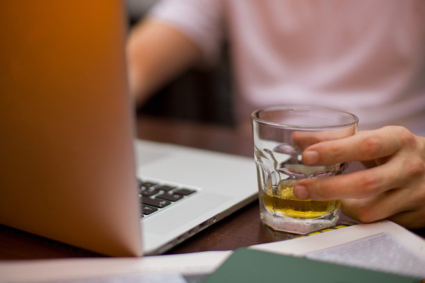 一个年轻人一边喝着威士忌，一边在笔记本电脑上远程工作，注意到他旁边的一部电话图片下载