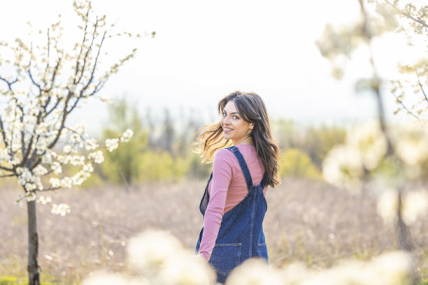美丽的姑娘，手里拿着草帽，站在春天盛开的梅枝上。春天的白花散了焦。图片下载