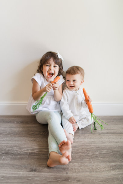 2023年春天，一个有着棕色卷发的4岁古巴裔美国女孩和她15个月大的穿着白色兔子装的小弟弟赤脚在家，在一个中性简单的空间里吃着一整根胡萝卜图片下载