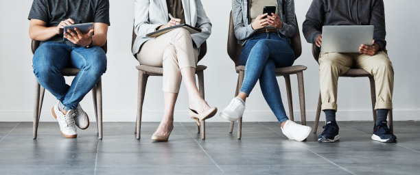 考生坐在一排的椅子上，使用科技设备。形形色色的应聘者等待面试，上网浏览面试问答。招聘机构招聘图片下载