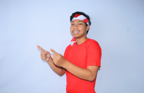 快乐的印尼年轻人穿着红色的t恤，戴着白色的国旗头巾，微笑着用手指着展示产品的一边图片下载