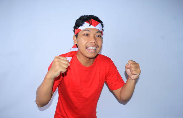 图为身穿红色t恤，头戴头巾的印尼青年在独立日高举拳头庆祝成功或进球。年轻人对自己的成就和满足做出“是”的手势图片下载