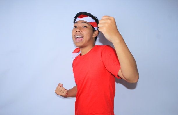 图为身穿红色t恤，头戴头巾的印尼青年在独立日高举拳头庆祝成功或进球。年轻人对自己的成就和满足做出“是”的手势图片下载