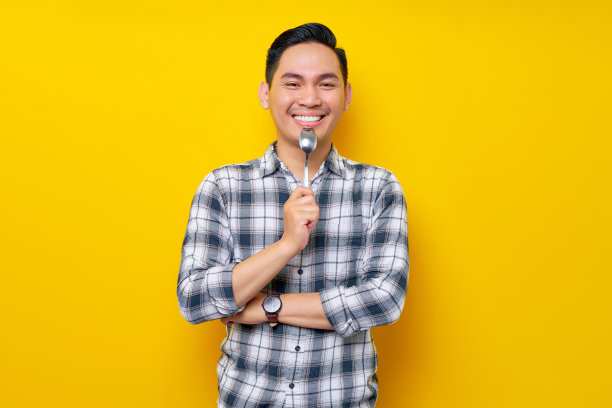 热情的年轻亚洲人穿着白色格子衬衫，拿着勺子品尝食物，孤立在黄色背景中。人的生活理念图片下载