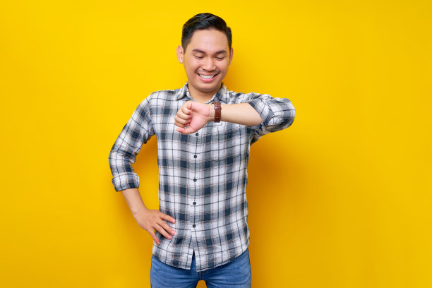微笑的年轻英俊的亚洲人穿着白色格子衬衫看着他的手表检查时间孤立的黄色背景。人的生活理念图片下载
