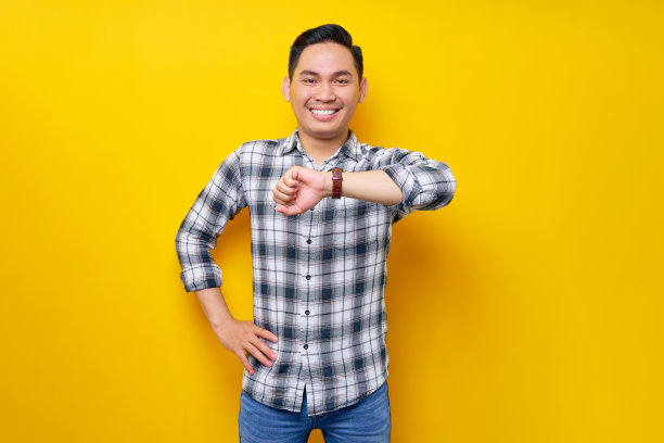微笑着快乐的年轻英俊的亚洲人穿着白色格子衬衫手表检查时间孤立在黄色背景。人的生活理念图片下载