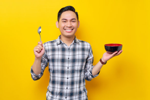微笑的年轻亚洲人穿着白色格子衬衫，手里拿着勺子和碗，准备品尝美味的食物，吃孤立的黄色背景。人的生活理念图片下载