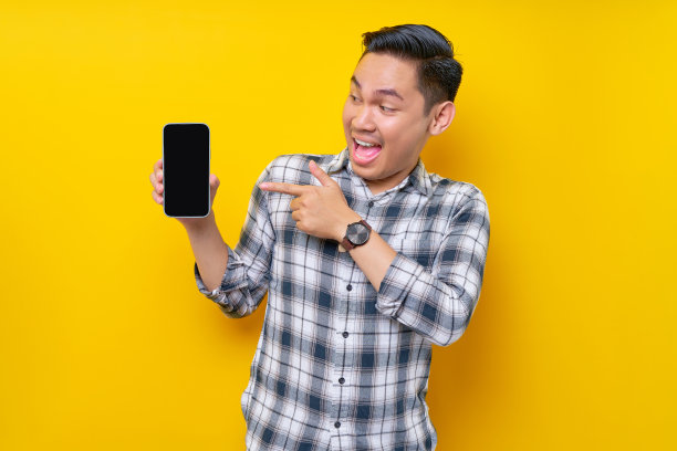 穿着白色格子衬衫的亚洲帅哥微笑着用手指着空白屏幕的手机，在黄色的背景上宣传新的手机应用程序。人的生活理念图片下载