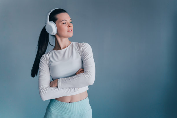 自信的黑发白人年轻女子穿着运动服，戴着耳机站在灰色的墙壁上，与复制空间望向别处。健康的生活方式，运动，健身。骄傲的美国女孩喜欢音乐。图片下载