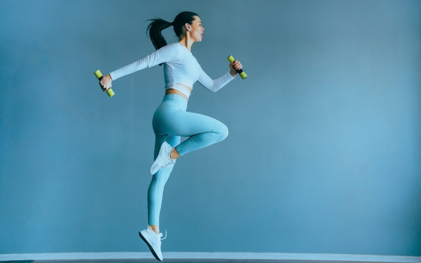 在健身课上，穿着蓝色运动服的健康白人年轻女子在普通的灰色墙壁上举着哑铃跳跃。高个黑发欧洲女孩扎着马尾辫做运动。健康的生活方式。图片下载