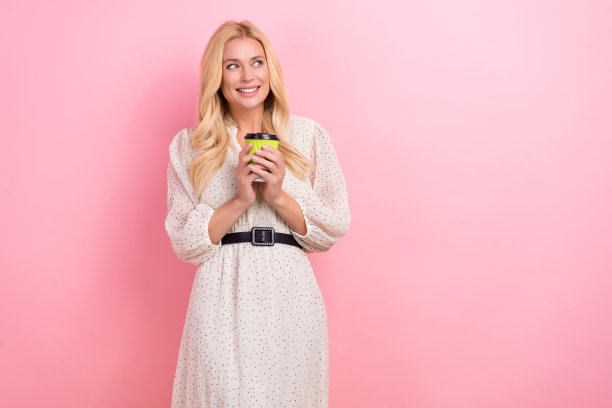 漂亮的金发女性的照片肖像拿着纸杯茶美式穿着时尚的白色服装孤立在粉红色的背景图片下载