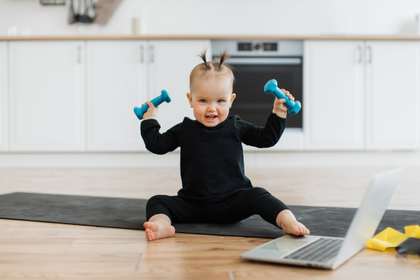 穿着运动服的婴儿在地板上的笔记本电脑旁举着哑铃图片下载
