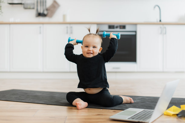 穿着运动服的婴儿在地板上的笔记本电脑旁举着哑铃图片下载