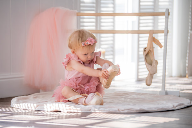 一个穿着粉色芭蕾舞服的可爱的小芭蕾舞女演员坐在房间里的扶手旁边，试图穿上她的足尖鞋。孩子和芭蕾舞。Copyspace。图片下载