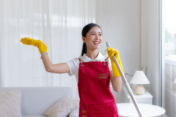 快乐的亚洲年轻女子工作拖地的房子唱歌拖把像在麦克风和玩得开心。戴着手套、系着围裙的亚洲主妇正在愉快地打扫房间。图片下载
