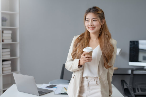成功的亚洲女商人在办公室拿着咖啡杯，用笔记本电脑微笑。自信的亚洲女商人愉快地坐在办公室里。图片下载