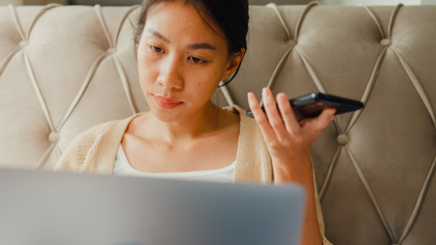特写:亚洲女孩把笔记本电脑放在床上，用智能手机向智能手机上的虚拟助手发出语音命令，早上在家里的卧室里录音。图片下载