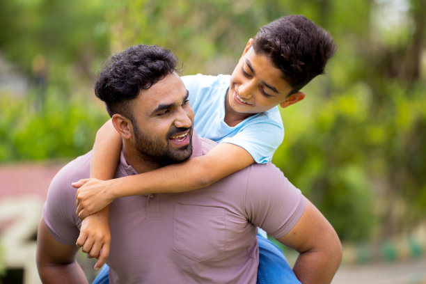 快乐兴奋的印度父亲背着他的小猪在公园里旋转儿子-童年记忆的概念，周末假期和无忧无虑的时刻。图片下载