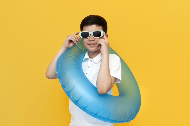 戴着太阳镜的亚洲小男孩拿着黄色背景的充气游泳圈，韩国孩子去度假图片下载