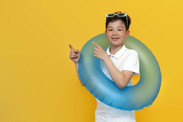 度假的亚洲小男孩拿着充气游泳圈，用手指着黄色背景的一侧图片下载