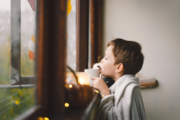 一个可爱的男孩裹在毯子里喝着热茶，看着窗外美丽的秋天图片下载
