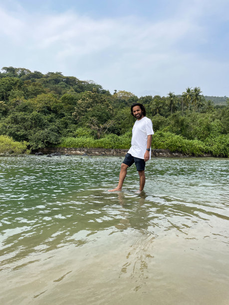 印度果阿帕洛莱姆，一名印度男子穿着短裤和白t恤，手拿凉鞋，光脚在水深及踝的海边划水，这是他的特写照片图片下载