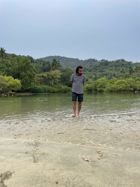 印度果阿，帕洛莱姆，一名印度男子穿着短裤和条纹t恤，手拿凉鞋，光脚在水深及踝的海边划水图片下载