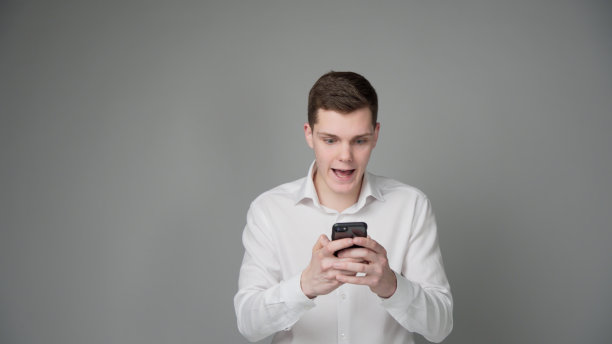 一个穿着白衬衫的年轻人正在热情地使用他的智能手机。他仔细看了看手机屏幕。使用手机应用程序或游戏。灰色的背景。图片下载