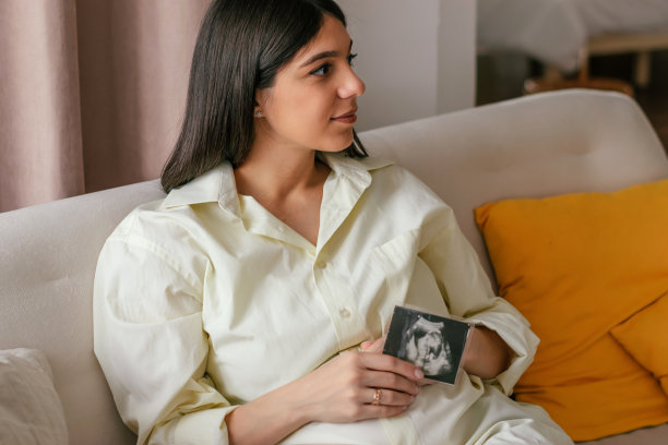 身穿黄色衬衫的年轻孕妇坐在家里的沙发上，手里拿着一张超声波照片。妊娠及产前护理理念。图片下载