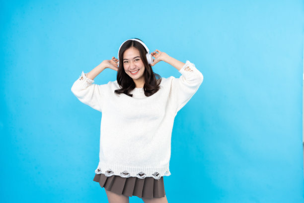 把白色的无线耳机戴在头上，微笑，享受听音乐的乐趣。穿一件外套和一条裙子。在摄影棚里拍一张蓝色的照片。图片下载