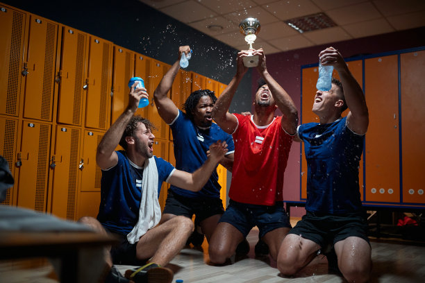 一群兴高采烈的年轻运动员在更衣室里庆祝，他们拿着金色的奖杯，拿着第一名。图片下载