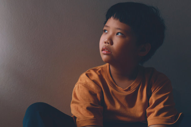 一个亚洲小男孩，带着悲伤和孤独的表情，躲在家里角落的阴影里，看着窗户，陷入了沉思图片下载
