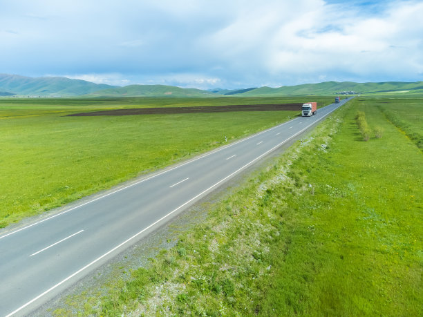 远处，红色的卡车沿着公路行驶，沿着绿色的田野和山脉，从无人机上看。从无人机上拍摄。货物交付和运输的概念图片下载