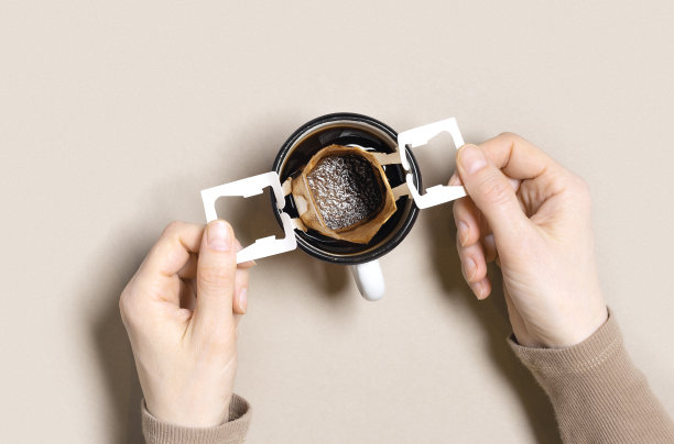 女人手握咖啡杯与黑咖啡饮料，滴滤咖啡袋与咖啡粉冲泡在杯子，纸袋滴滤咖啡，米色背景，俯视图，最小的平面，柔和的颜色图片下载