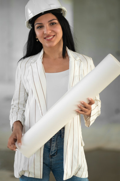 戴着安全帽的年轻女建筑师面带微笑，手里拿着图纸，看着镜头。施工经理，经理，施工现场的工程师。垂直图图片下载