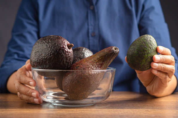 一只手拿着一个新鲜的牛油果，三只牛油果放在木桌上的一个玻璃碗里图片下载