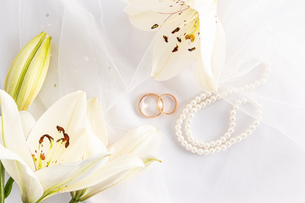 俯视图两个金色的结婚戒指在一个白色的新娘新娘面纱和珍珠珠。精致的白色百合花。婚礼贺卡的概念，图片下载