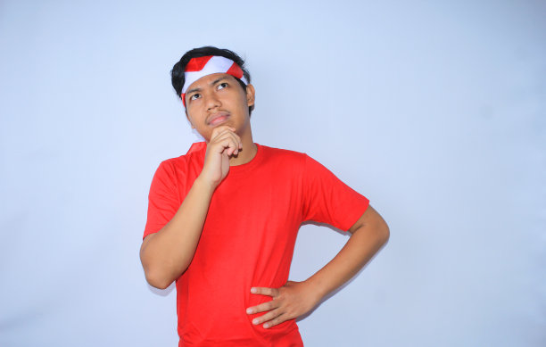 印尼独立纪念日，一名穿着红色t恤，头戴头巾，双手捂着下巴，思考和怀疑的印尼青年孤立白色图片下载