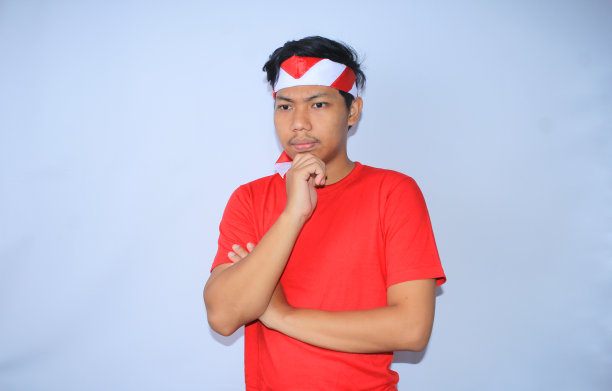 印尼独立纪念日，一名穿着红色t恤，头戴头巾，双手捂着下巴，思考和怀疑的印尼青年孤立白色图片下载