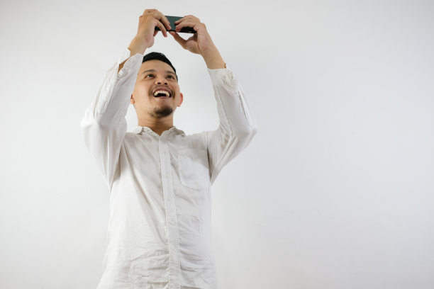 一个穿着休闲衬衫的迷人亚洲男人的肖像，他通过倾斜手机屏幕玩游戏。开心的表情。白色背景上的孤立图像图片下载