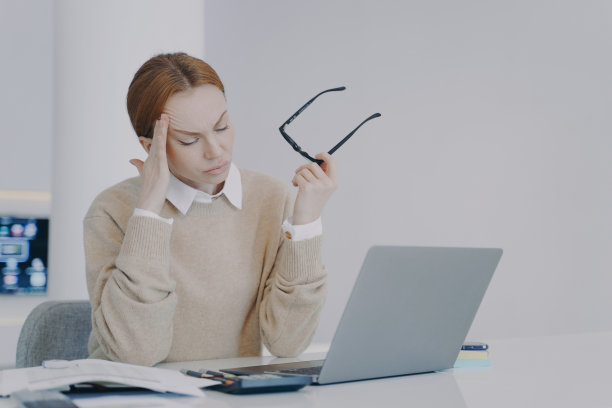一位疲惫不堪的年轻女士坐在办公室的办公桌前，摘下了眼镜。员工在工作场所头痛。图片下载