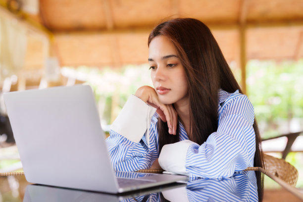 照片中，亚洲女人在桌子前放着一台笔记本电脑，脸上有纠正错误的严肃表情，假期里有应该休息的无聊表情，却有坐着工作。图片下载