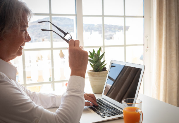成熟的资深白发女性使用笔记本电脑键盘打字，聪明的工作理念。一位老妇人坐在窗前的桌子旁图片下载