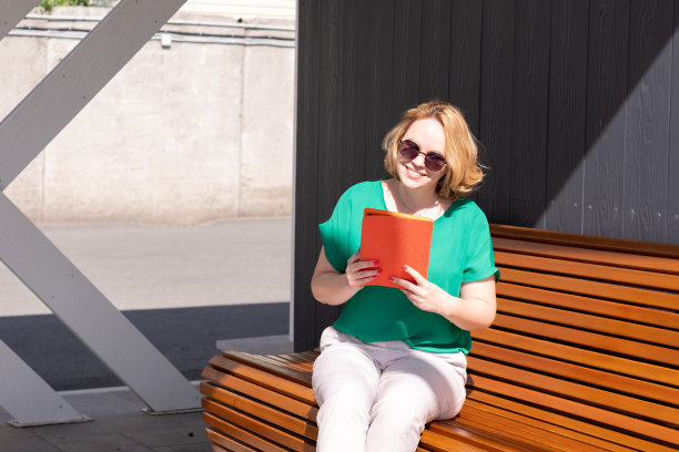 一位戴着墨镜的微笑女子手持平板电脑，在街上的长椅上阅读新闻，使用应用程序。图片下载