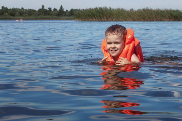 一个微笑的小男孩穿着橙色的救生衣在蓝色的湖水中游泳。图片下载