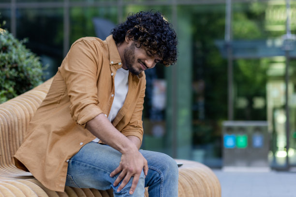 一个年轻的印度男子坐在外面的长凳上，抱着膝盖。腿部受伤，疼痛难忍，正在等待紧急医疗救助图片下载