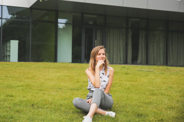 美丽的时髦女孩在阳光明媚的日子里坐在现代建筑附近。时尚的商务女性在绿色的草坪上休息。学生的生活自由时间。上班族休息。图片下载